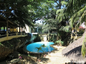 Ein Pool, umgeben von tropischer Vegetation, einer strohgedeckten Struktur und einer Steinmauer.