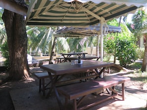 A covered picnic area with wooden tables and benches, surrounded by palm trees and greenery.