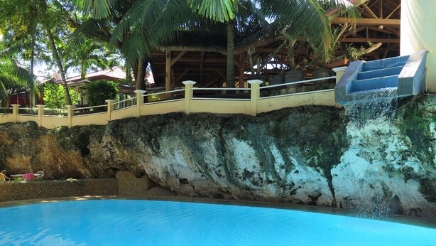 Una piscina con cascada, rodeada de un muro de piedra y palmeras.