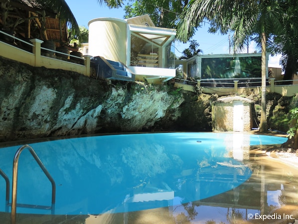 A swimming pool with a diving platform, surrounded by a rocky cliff and palm trees.