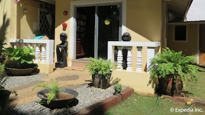 Un edificio amarillo con un balcón blanco, una puerta y un pequeño jardín con plantas y una escultura decorativa.