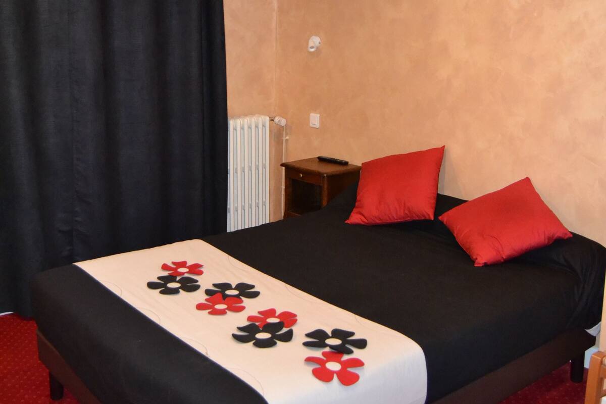 Un dormitorio con cama, almohadas negras y rojas, colcha blanca y negra, alfombra roja, cortina negra, mesita de noche de madera y pared blanca.