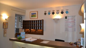 Een hotel receptie met een houten balie, ingelijste kunstwerken, klokken die verschillende steden weergeven, en een menukaart met prijzen voor kamers.