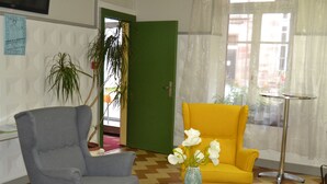 Een kamer met een grijze fauteuil, een gele fauteuil, een kleine tafel met een bloemenvaas, een groene deur en een raam met dunne gordijnen.