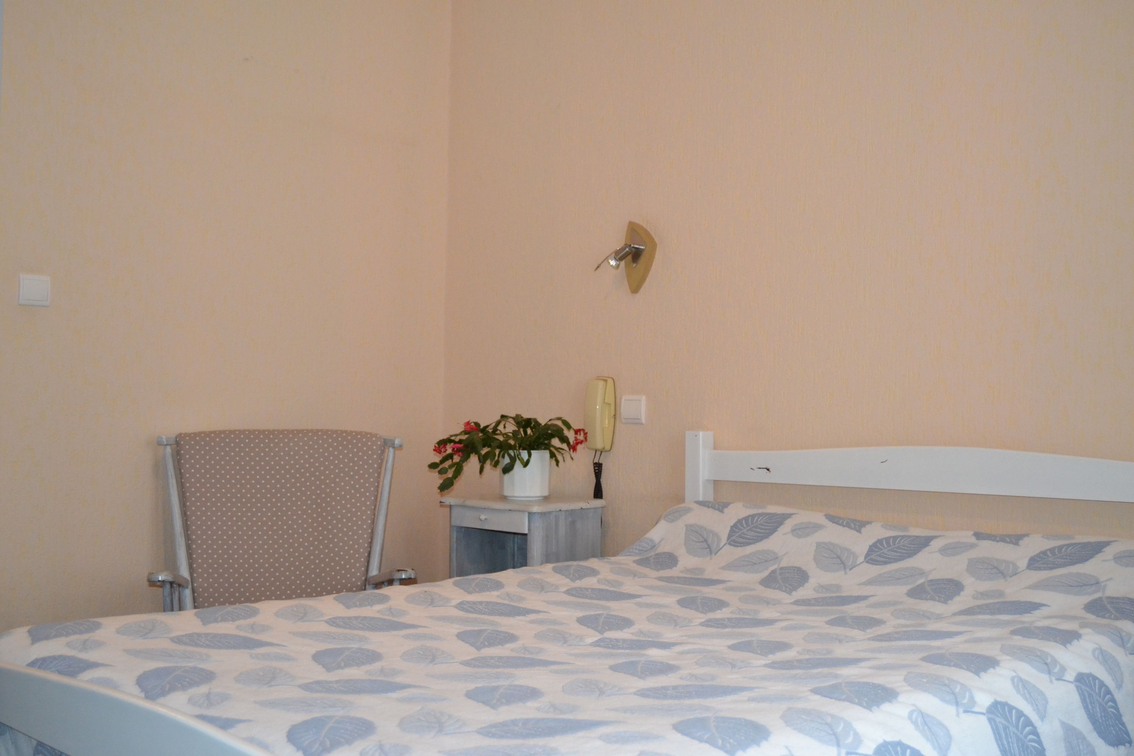 A bedroom with a bed, a chair, a small table with a plant, and a wall-mounted light fixture.