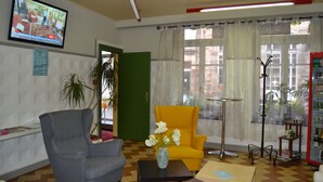 Een woonkamer met een televisie, een gele fauteuil, een grijze fauteuil, een klein tafeltje met een vaas en een vloerlamp.