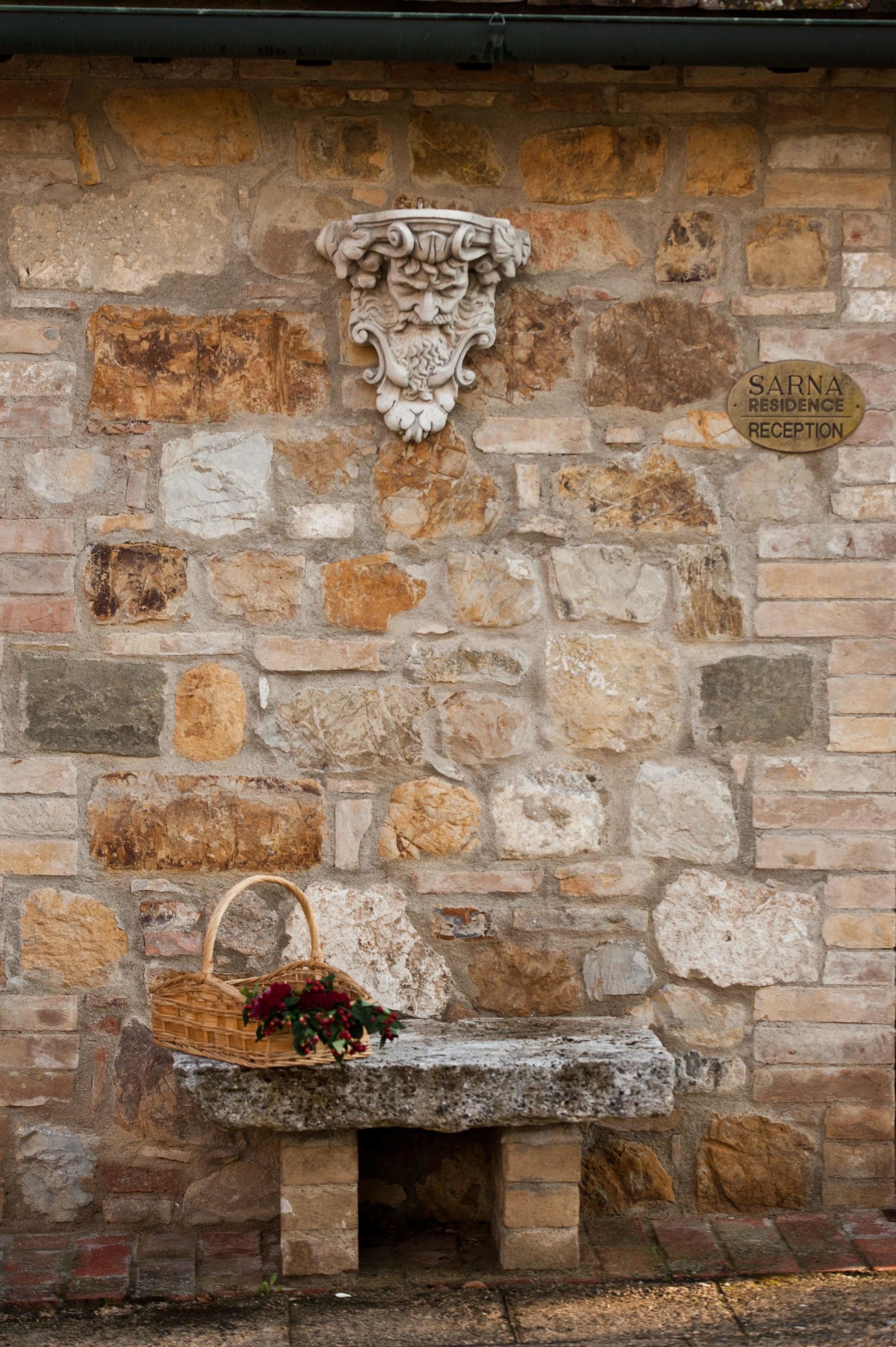 Un muro de piedra con un banco de piedra y una cesta con flores. Un letrero dice 