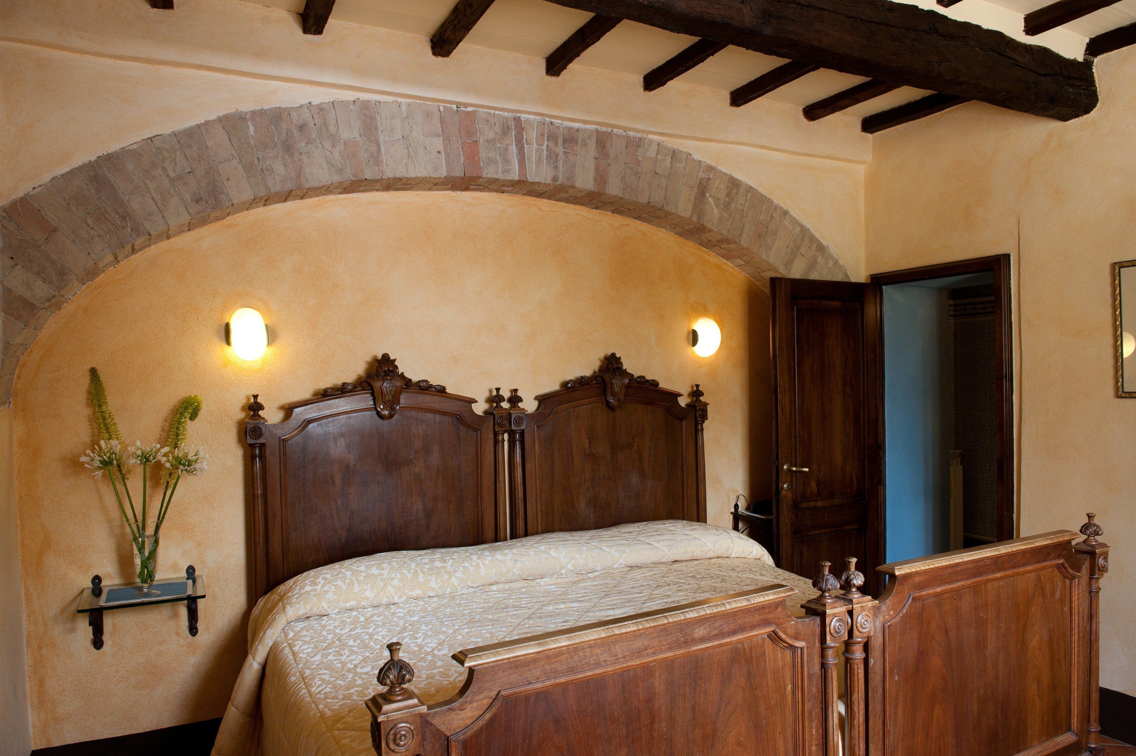 A bedroom with a wooden bed, an arched stone wall, and two wall-mounted lights.