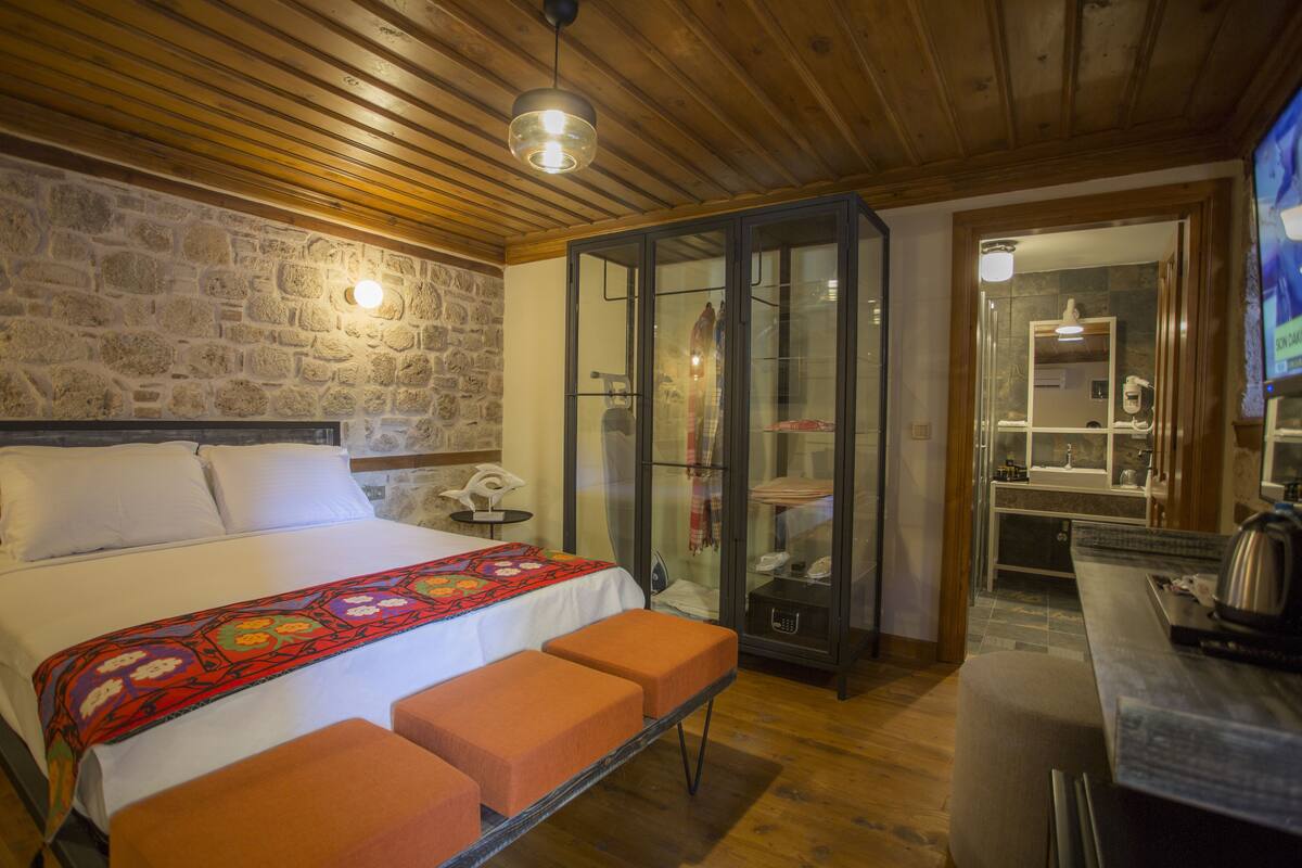 Ein Schlafzimmer mit Steinwand, einem Bett mit bunter Decke, Holzboden und einem Glaskabinet.