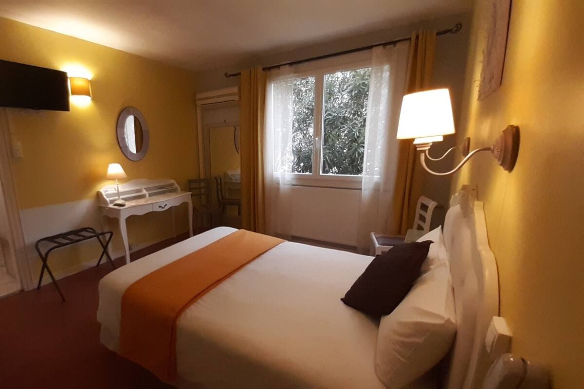 A hotel room with a bed, a desk, a chair, a TV mounted on the wall, and a window with curtains.