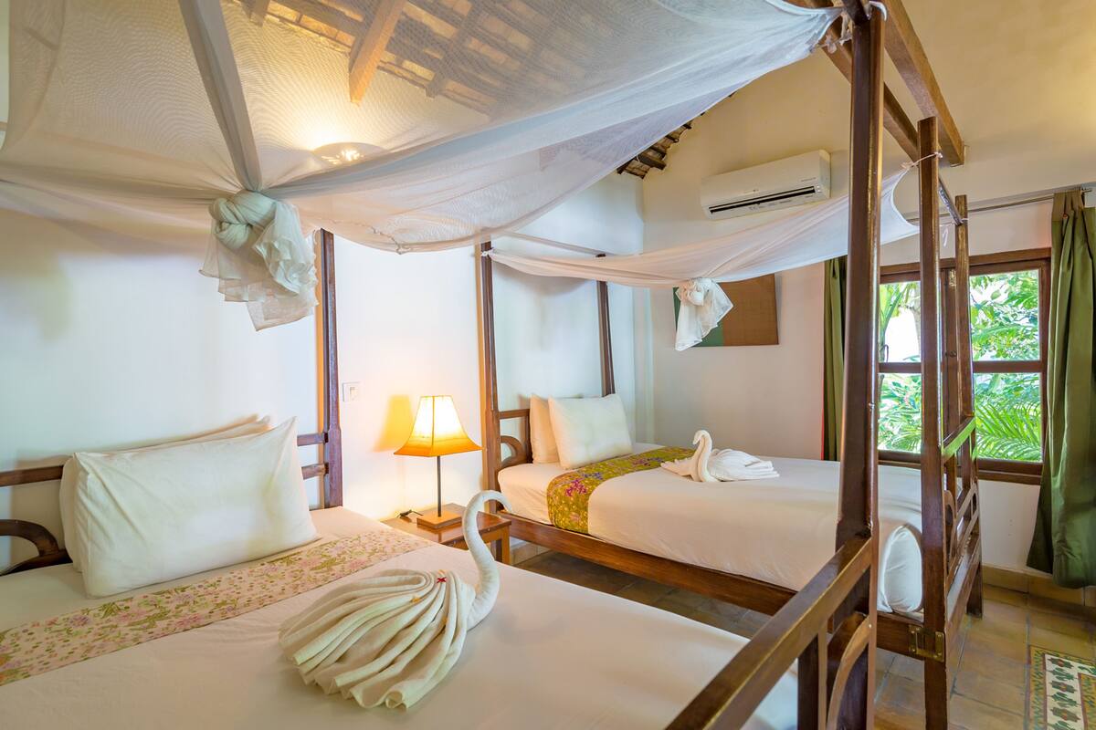 Quarto de hotel com duas camas, incluindo uma cama de dossel, mesa de cabeceira com abajur e janela com cortinas.