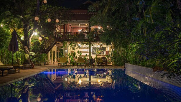 Zona junto a la piscina con comedor, rodeada de abundante vegetación y un edificio con balcón.