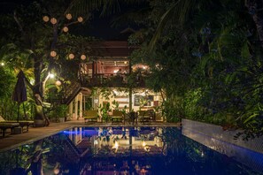 Zona junto a la piscina con comedor, rodeada de abundante vegetación y un edificio con balcón.