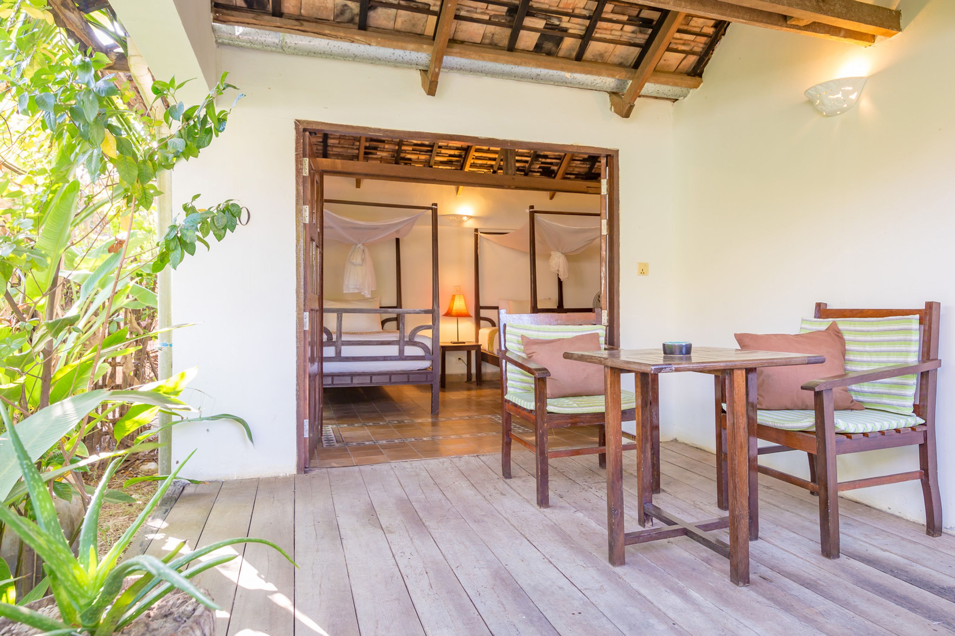 A wooden deck with a table and chairs, a bench, and a doorway leading to a room with a bed and a lamp.