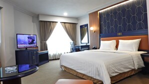 Ein Hotelzimmer mit einem großen Bett, einem Schreibtisch mit Fernseher, einem Nachttisch und einem Vorhangfenster.