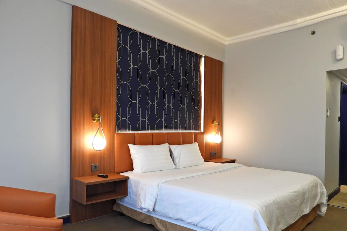 A hotel room with a bed, wooden headboard, two bedside tables, a nightstand, a chair, and a wall-mounted light fixture.
