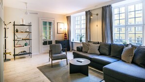 A modern living room with a sofa, armchair, and coffee table. There is a bookshelf, a door, and a window with curtains. - Churchill 3BR by Daniel&Jacob's (Copenhagen)