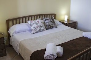 A neatly made bed with white bedding and two patterned pillows, a wooden headboard, a bedside table with a lamp, and a rolled towel on the bed. - Residence Villa Santa Lucia (Cefalù)