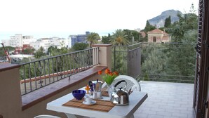 A balcony with a table set for two, a chair, and a view of the city and mountains. - Residence Villa Santa Lucia (Cefalù)