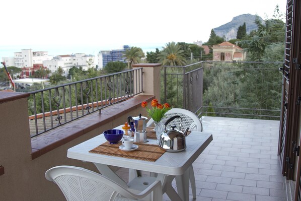 A balcony with a table set for two, a chair, and a view of the city and mountains. - Residence Villa Santa Lucia (Cefalù)