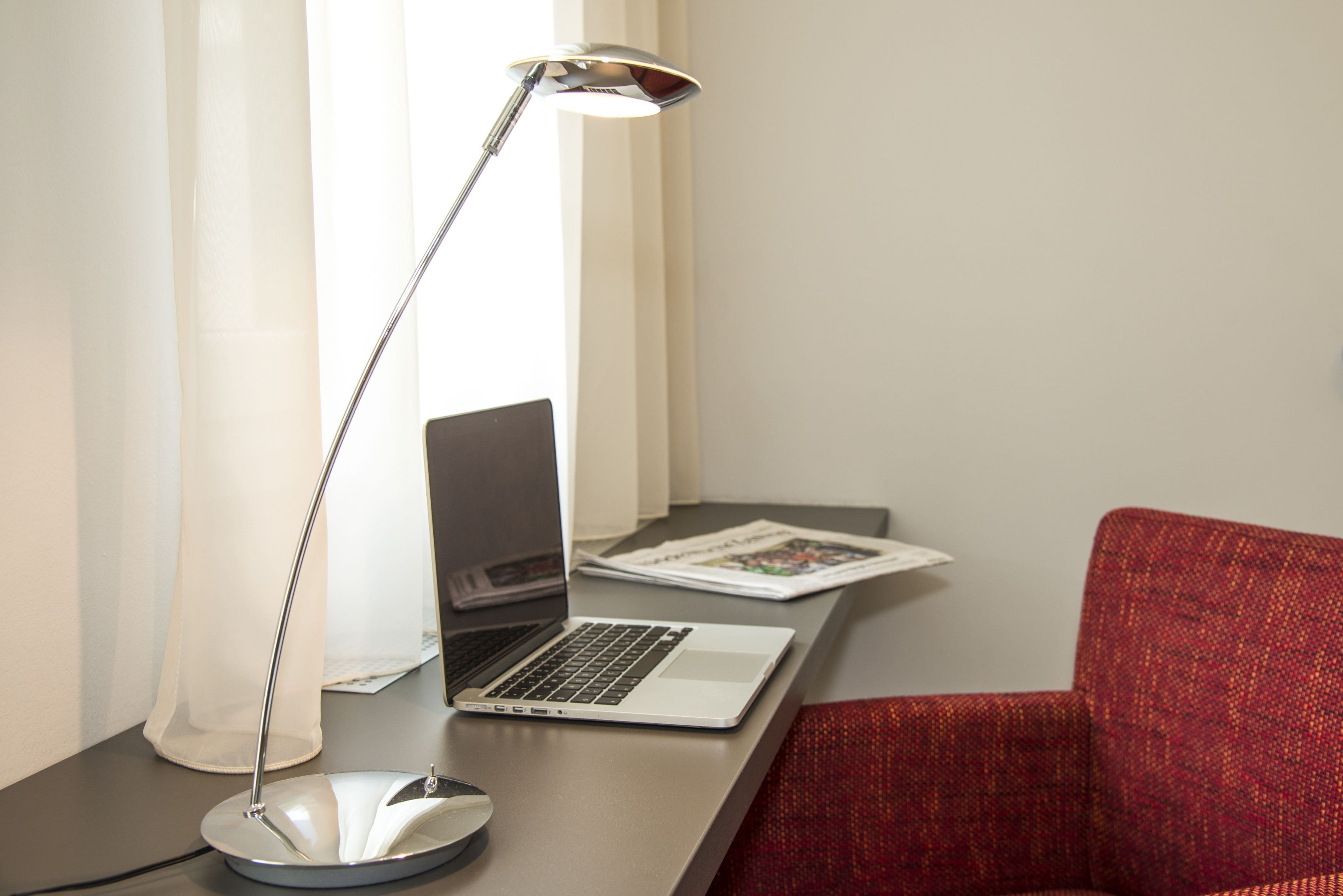 En bærbar computer på et skrivebord med en skrivebordslampe, en rød stol og et magasin.