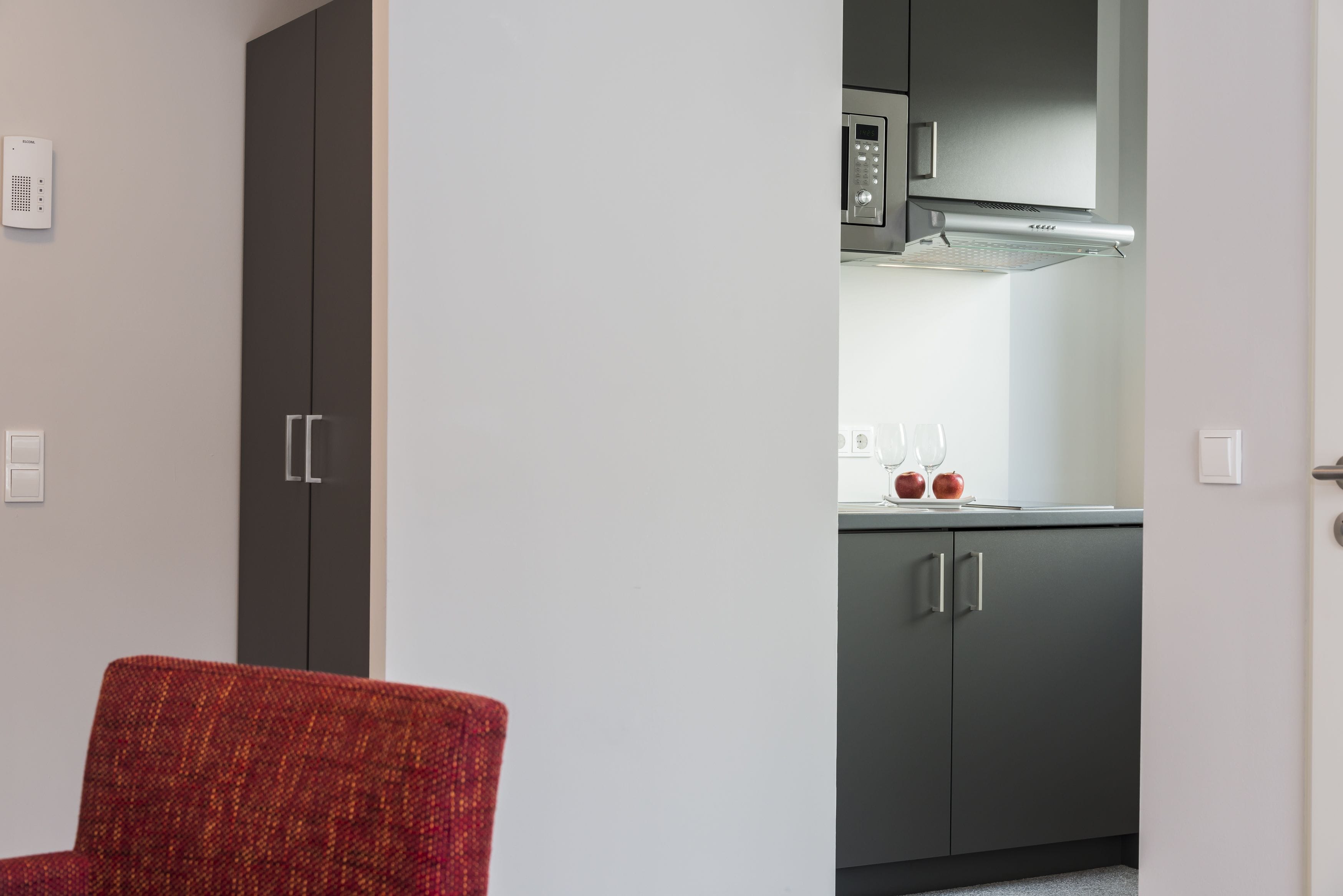 A compact kitchen with grey cabinets, a microwave, and a red chair.
