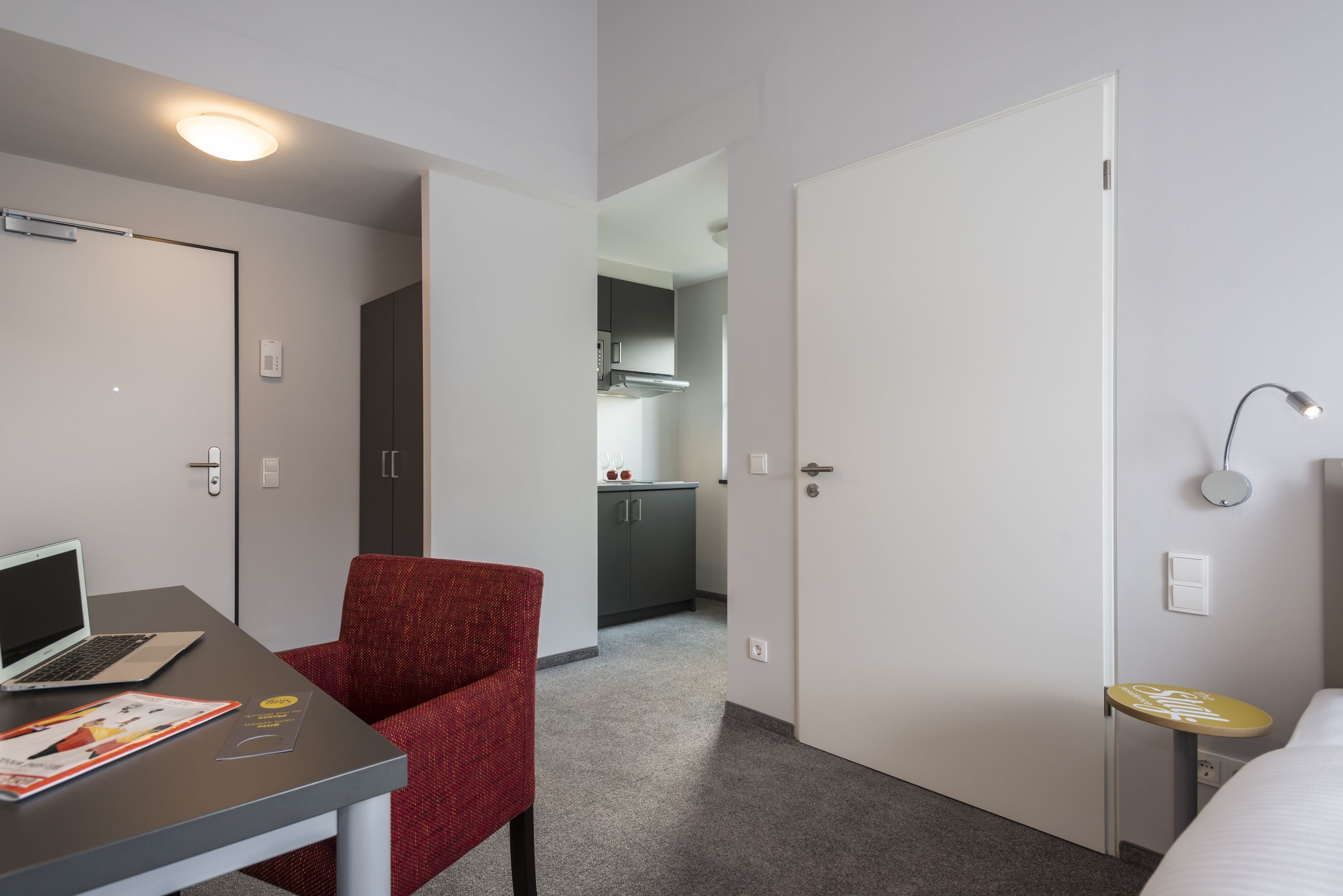 Quarto de hotel com escrivaninha, cadeira, cama e uma mesinha com abajur.