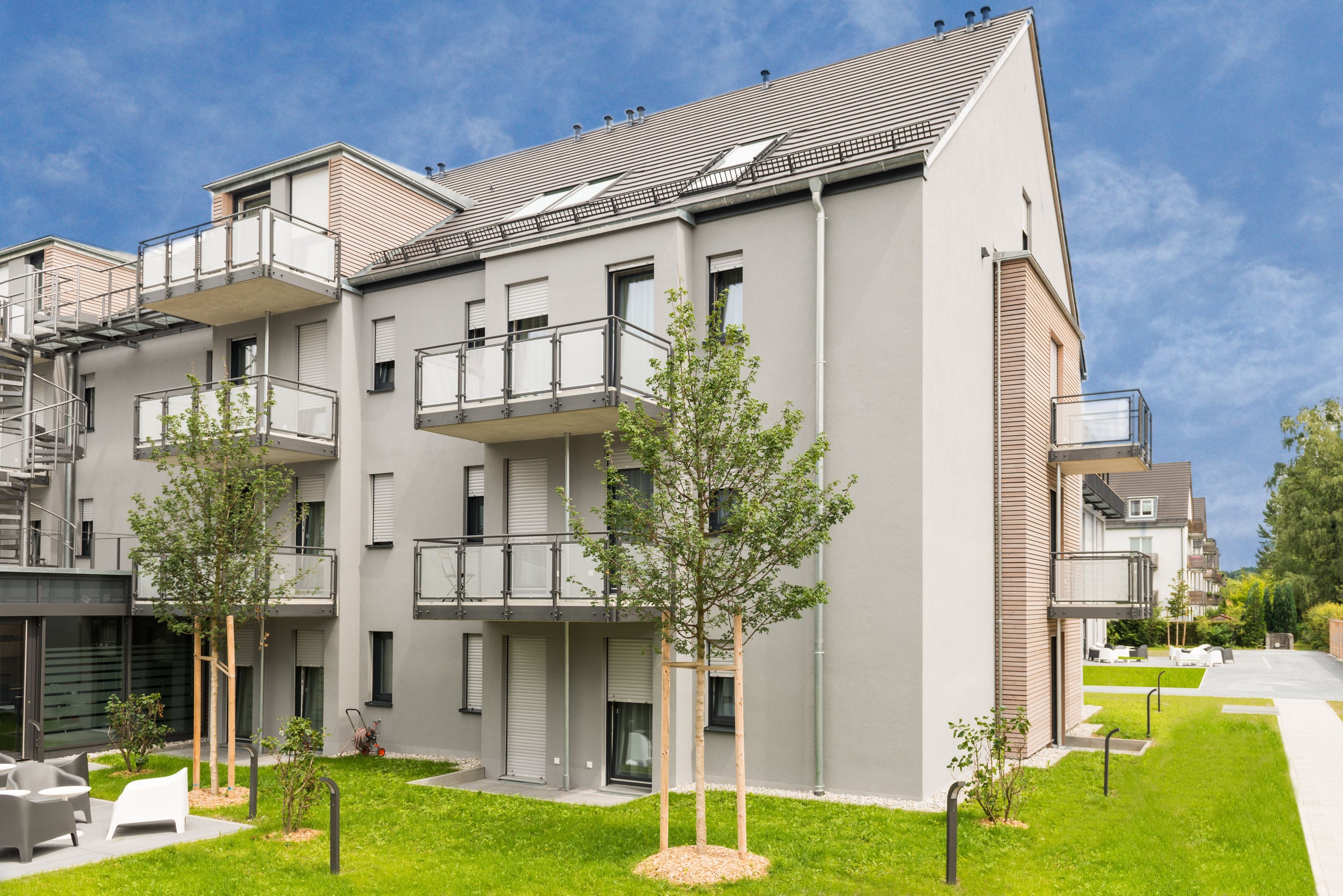 A modern multi-story residential building with balconies, a small courtyard, and a well-maintained lawn.