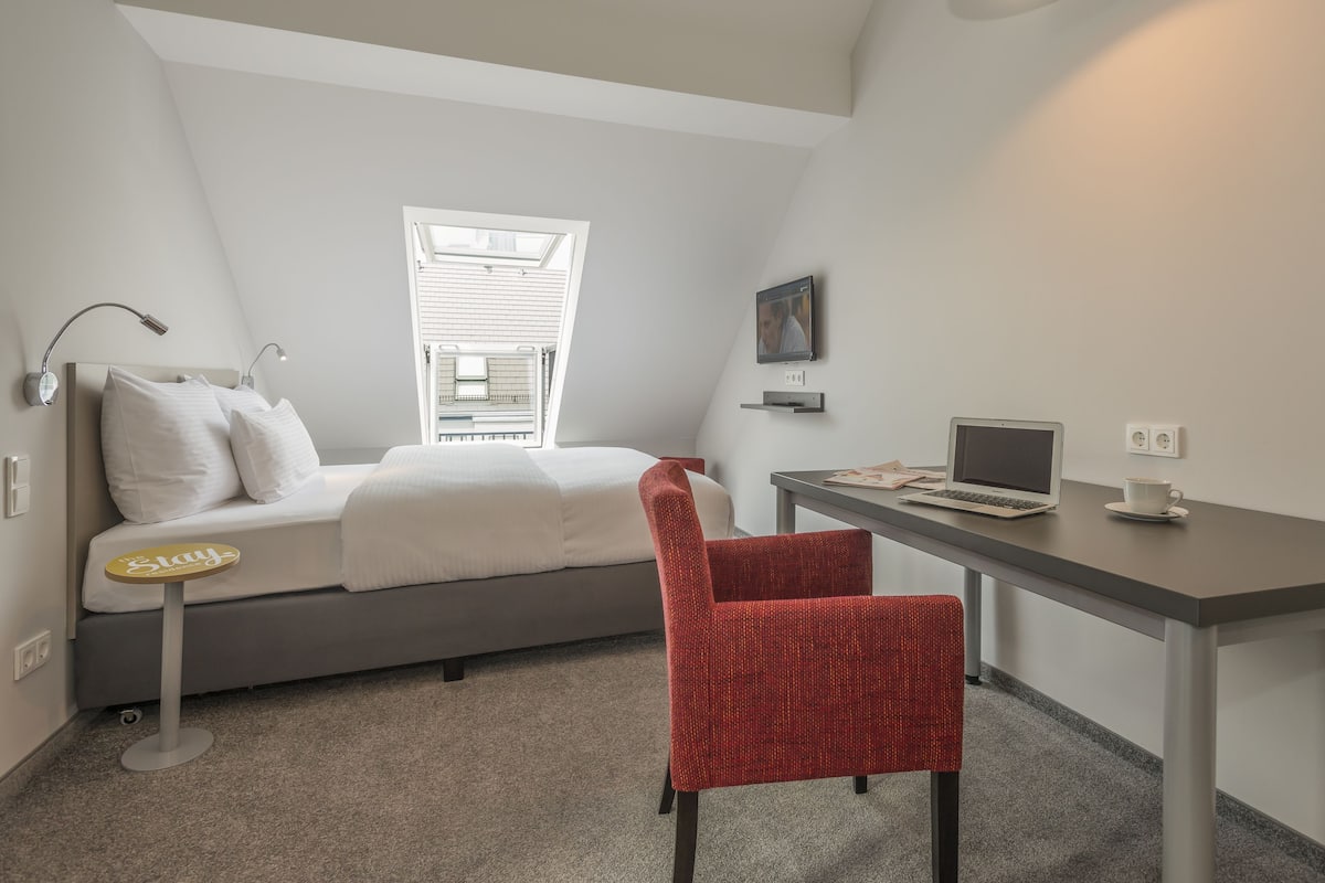 A bedroom with a bed, a desk, a chair, a laptop, and a window.
