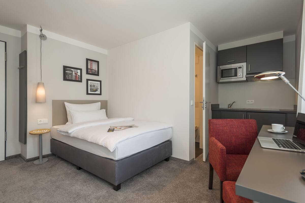 A hotel room with a bed, a desk with a laptop, a red chair, a microwave, and a small table with a cup.