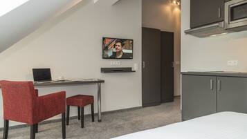 A modern, compact room with a bed, a desk with a laptop, a television, and a kitchenette.