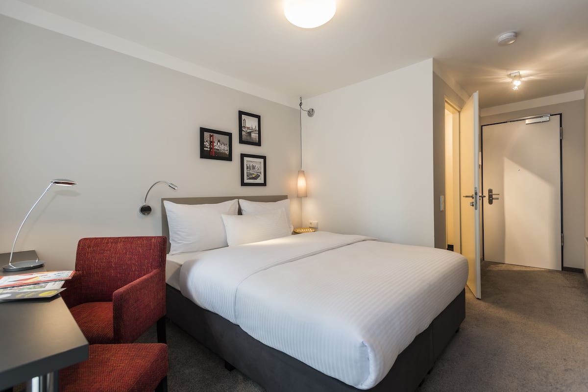 A hotel room with a bed, a desk with a chair, a lamp, and framed pictures on the wall.