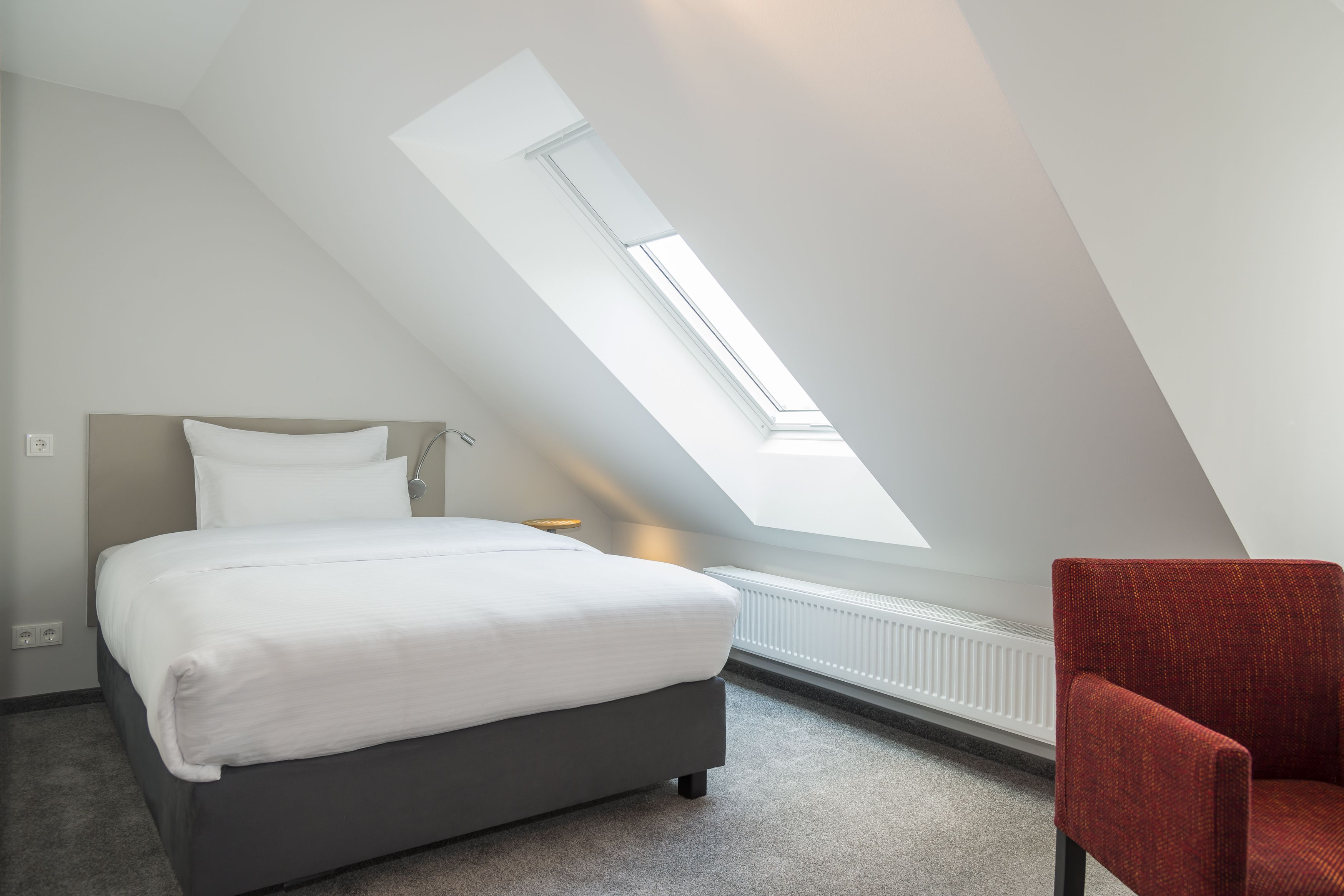 Een slaapkamer met dakraam, een bed, een rode stoel en een radiator.