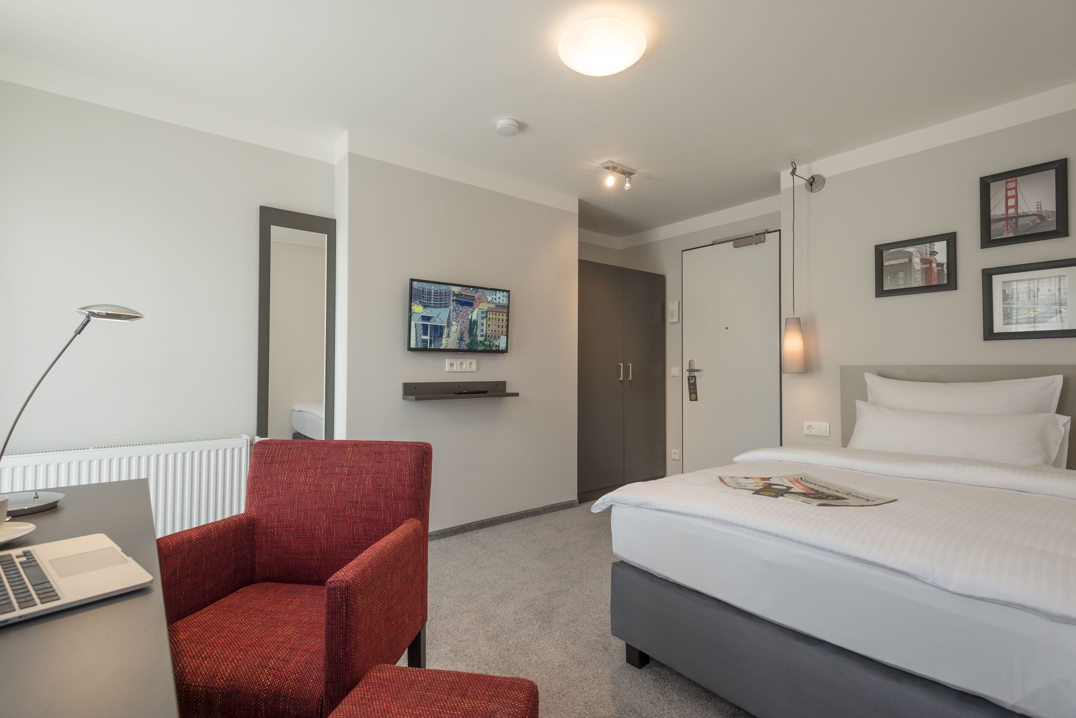 Una habitación de hotel con una cama, un escritorio con una computadora portátil, una silla roja, un televisor en la pared y una puerta que da a otra habitación.