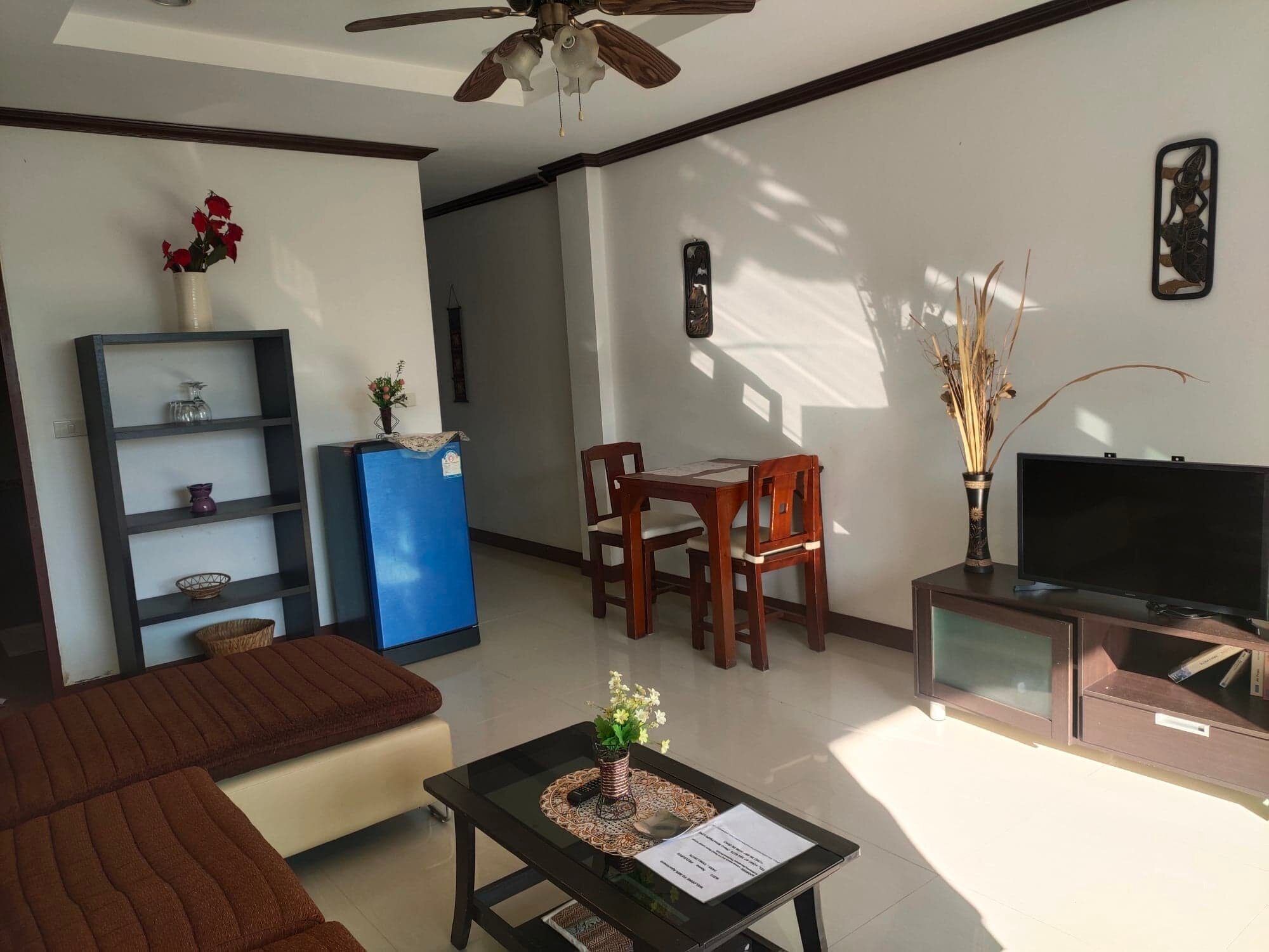 Una sala de estar con un sofá, una mesa de centro con un florero, un refrigerador azul, una mesa de comedor de madera con sillas, un televisor sobre un mueble bajo y un adorno en la pared.