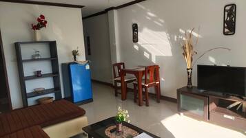 Una sala de estar con un sofá, una mesa de centro con un florero, un refrigerador azul, una mesa de comedor de madera con sillas, un televisor sobre un mueble bajo y un adorno en la pared.