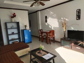 A living room with a sofa, a coffee table with a vase, a blue refrigerator, a wooden dining table with chairs, a television on a stand, and a decorative wall piece. - BEA Apartment (Koh Samui)