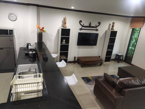 A modern living room with a kitchen area, a flat-screen TV mounted on the wall, and a brown leather sofa. - BEA Apartment (Koh Samui)