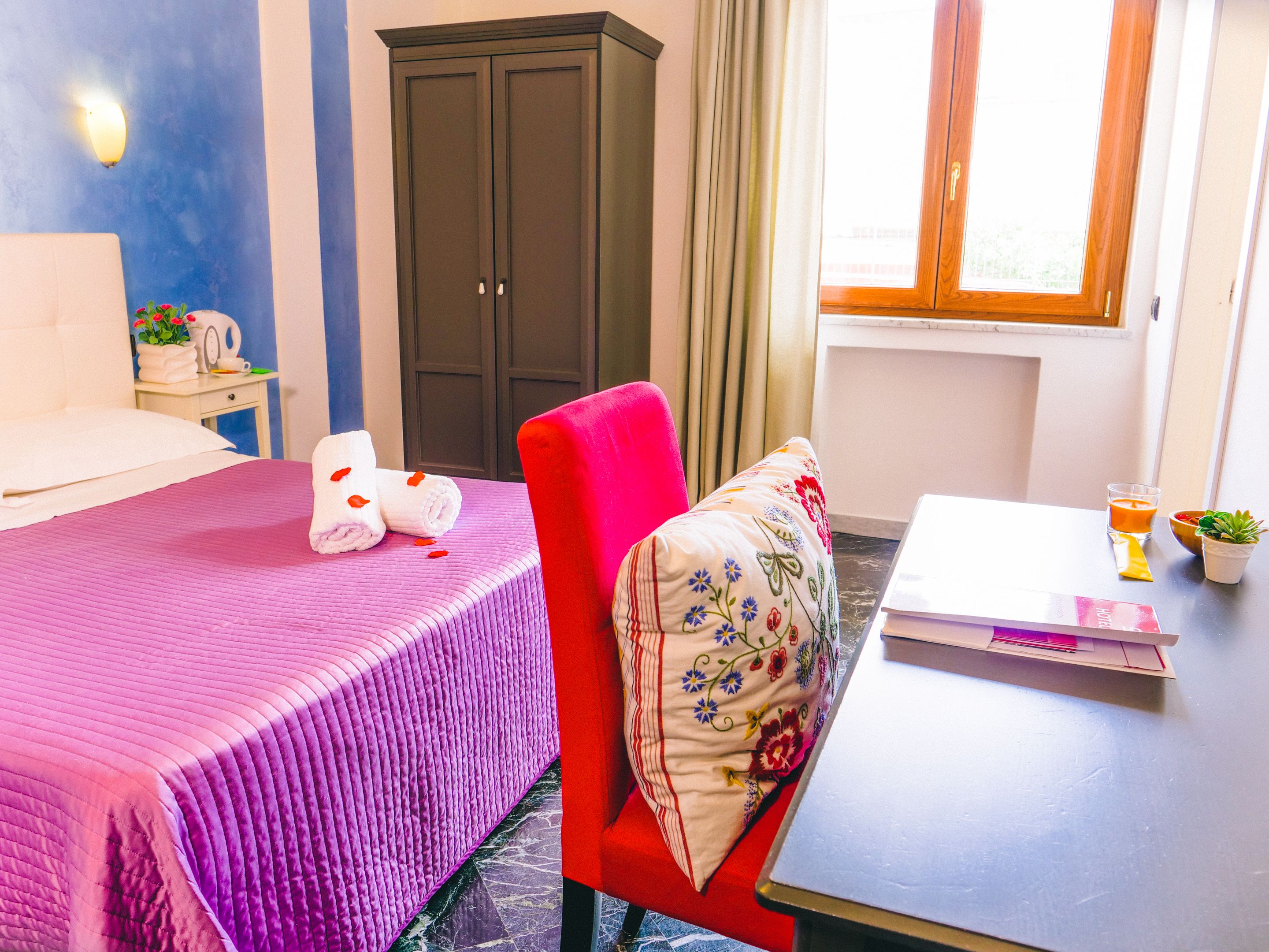Una habitación de hotel con una cama, una silla roja, una mesita con un vaso de zumo de naranja, un frutero y una planta en maceta.