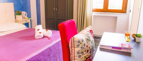 Una habitación de hotel con una cama, una silla roja, una mesita con un vaso de zumo de naranja, un frutero y una planta en maceta.