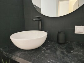Een moderne badkamer met een witte wastafel, donkere betegelde wanden en een marmeren aanrechtblad.
