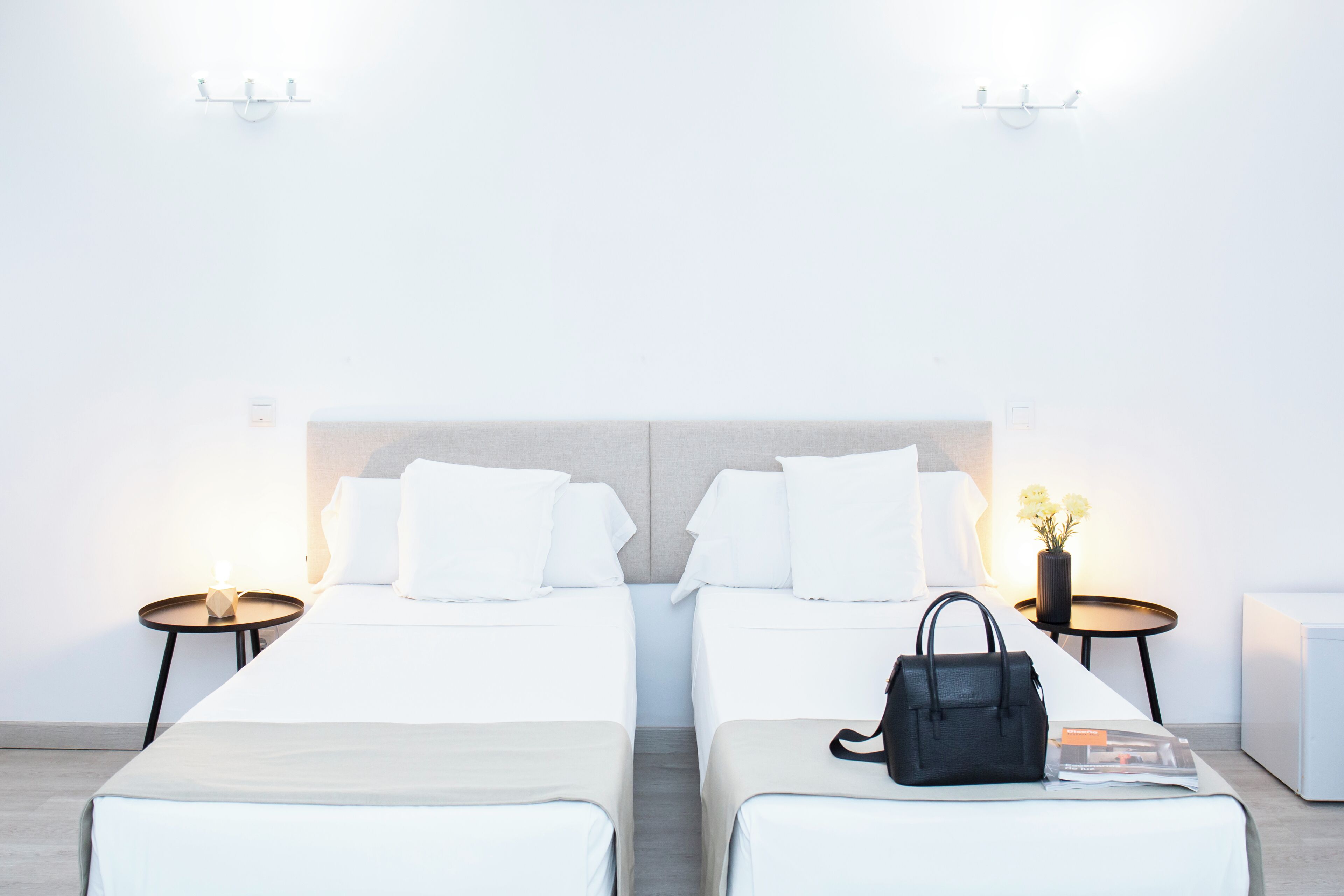 A hotel room with two beds, a nightstand with a lamp, a small table with a vase of flowers, and a black handbag on the bed.