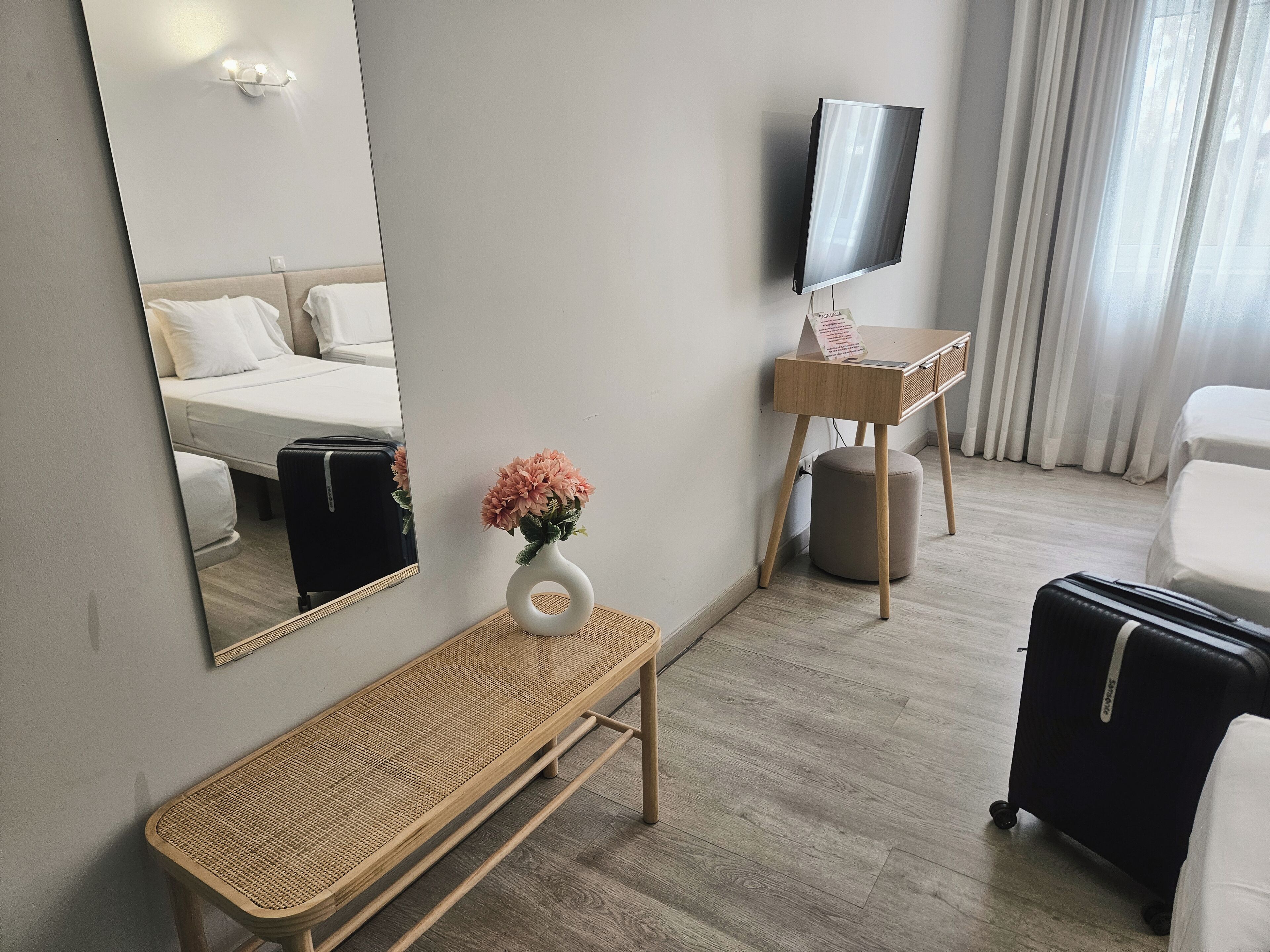 A hotel room with a bed, a desk with a TV, a bench, and a suitcase.