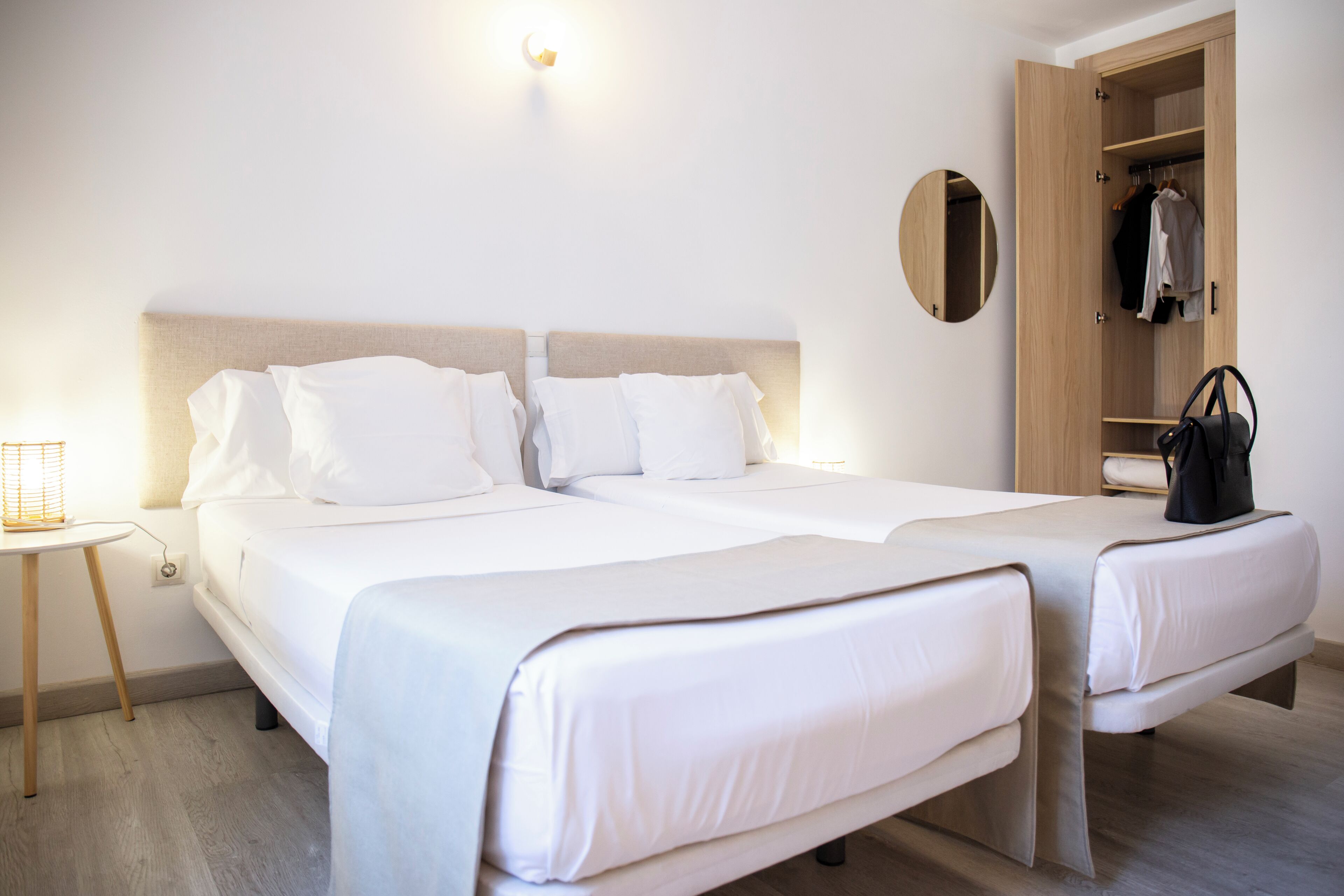 A double bed with white linens, a bedside table, a wooden wardrobe, and a round mirror.