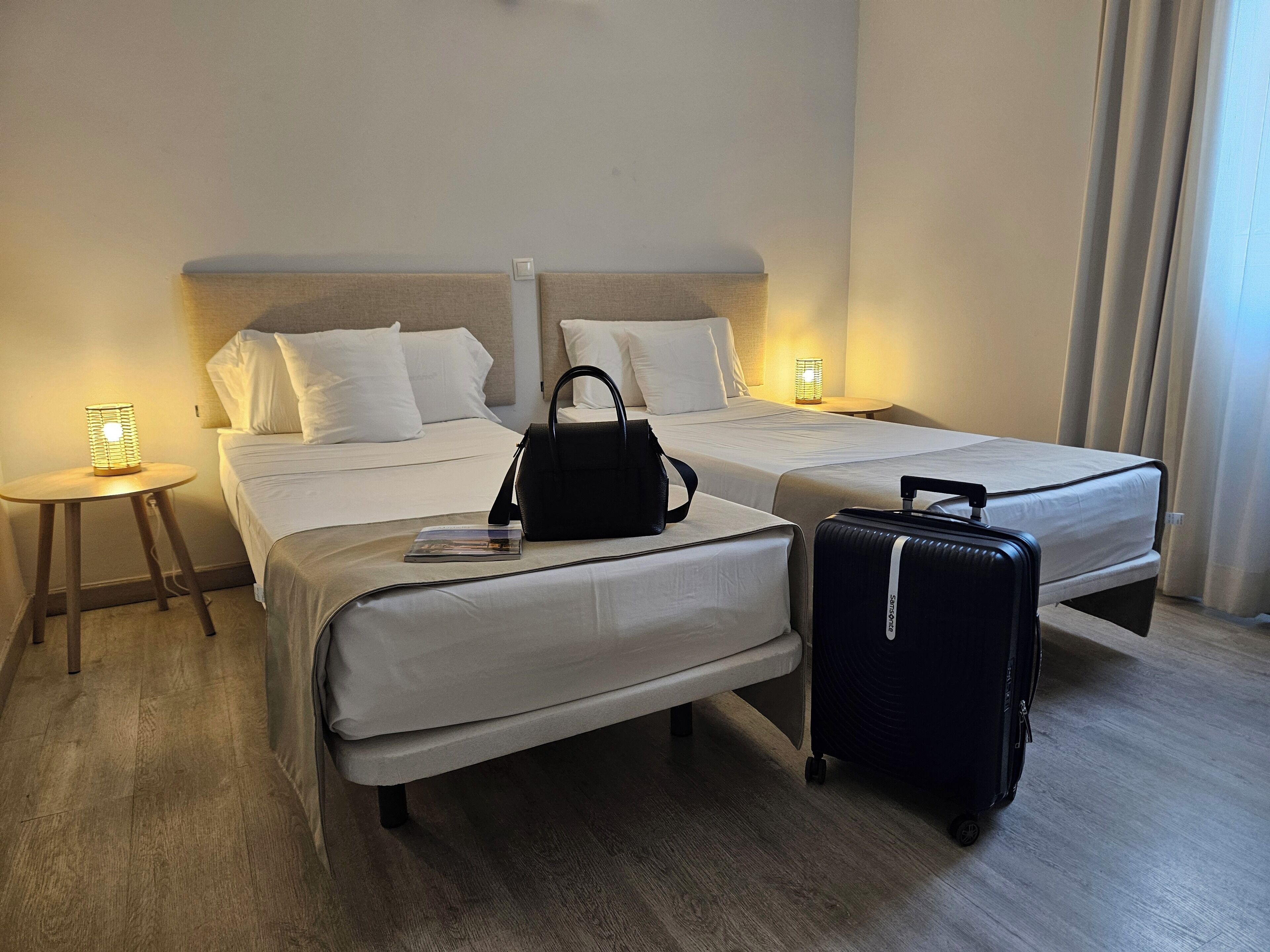 Ein Hotelzimmer mit zwei Betten, einem Nachttisch, einer Lampe, einer schwarzen Handtasche und einem Koffer.