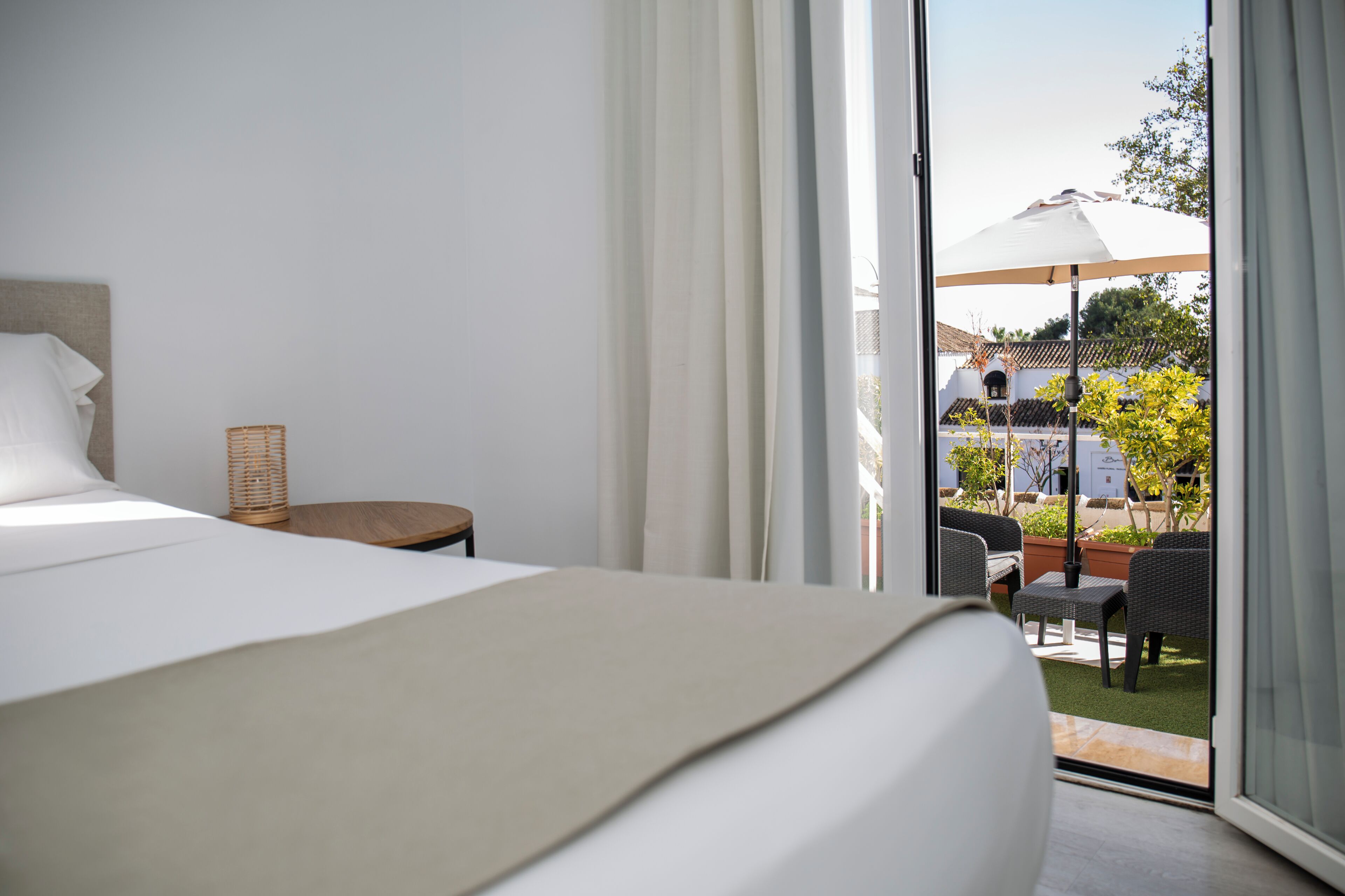 Una camera da letto con un letto, un tavolino e una sedia in vimini. Si vede un patio esterno con sedie e un ombrellone.