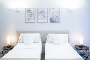 Een tweepersoonsbed met wit beddengoed, twee bedlampjes en drie florale kunstwerken in lijsten aan de muur.