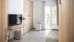 Habitación de hotel moderna con televisión de pantalla plana, una mesita con un florero, un refrigerador compacto, una cama y un balcón con vistas a la vegetación.