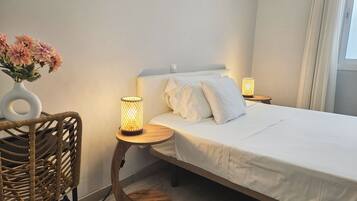 A neatly arranged bedroom with a bed, two bedside lamps, a wicker chair, and a vase with flowers.