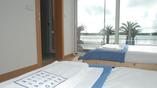 Ein Hotelzimmer mit großem Fenster und Blick auf ein Gewässer sowie Palmen. Es gibt zwei Betten mit Handtüchern, eines mit blauem und das andere mit weißem Handtuch.